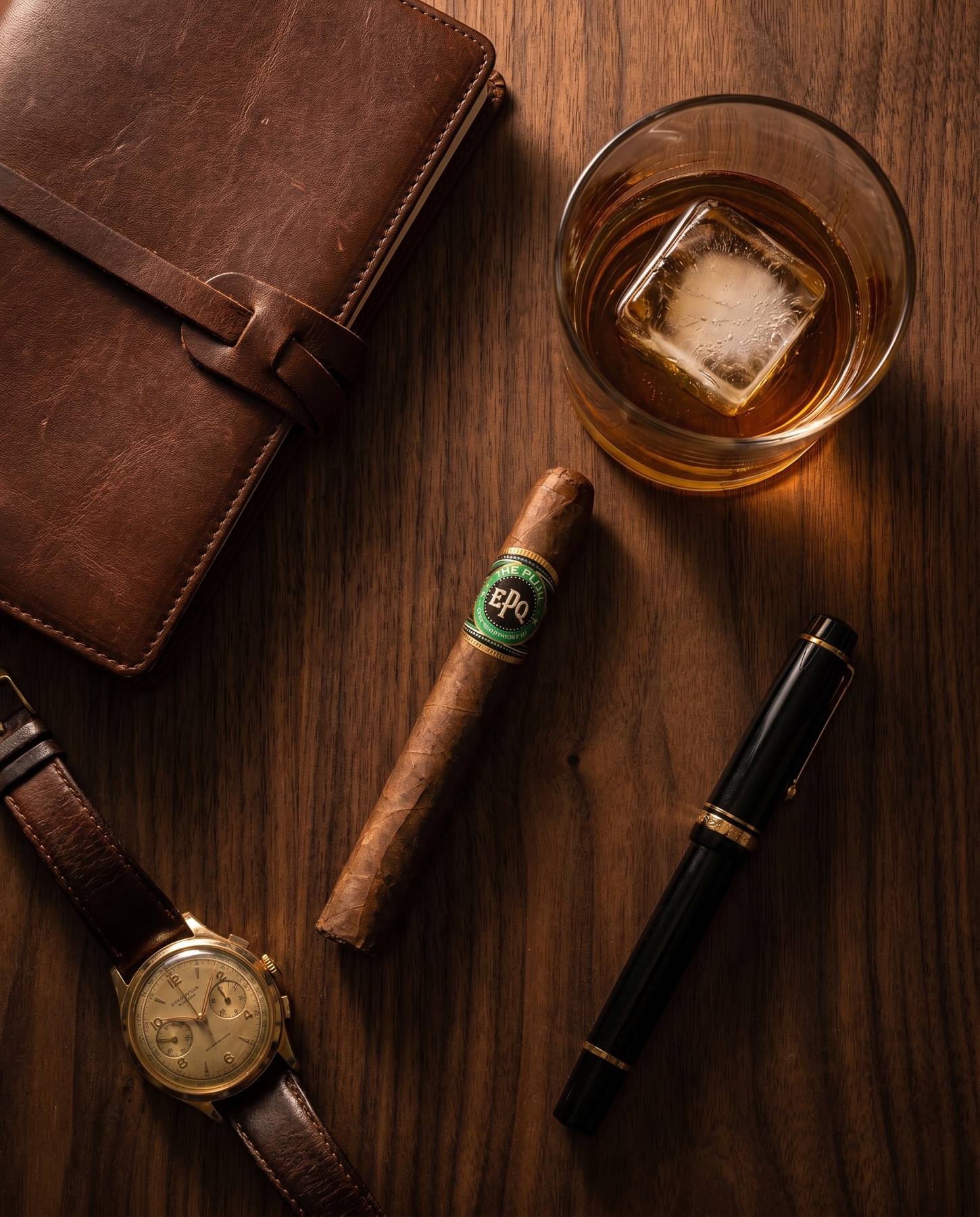 The Plug beside a leather journal, whisky, watch, and fountain pen on walnut